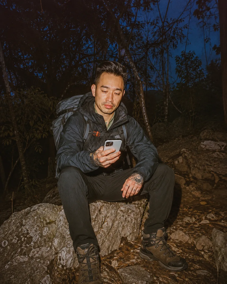 A hiker sitting out on a rock at dusk with a pack beside him, phone in both hands
