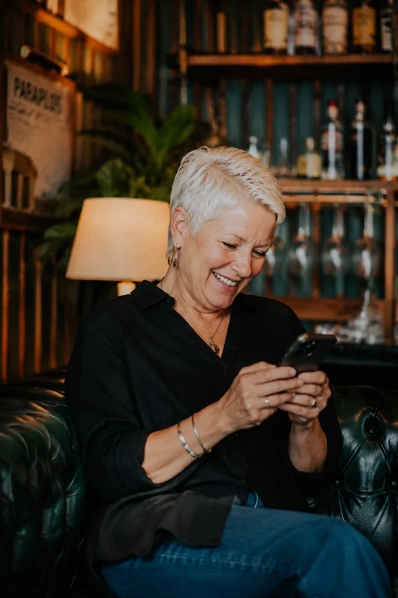 A woman in her fifties laughing at her phone in an armchair, a lamp lit behind her